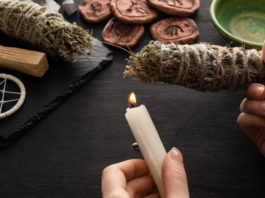 Cropped view of shaman holding candle and smudge stick near witchcraft on black wooden background