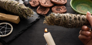 Cropped view of shaman holding candle and smudge stick near witchcraft on black wooden background