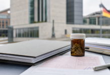 El seguro público alemán excluye la flor de cannabis medicinal Documentación sanitaria y un frasco neutro de cannabis medicinal ante un edificio institucional alemán