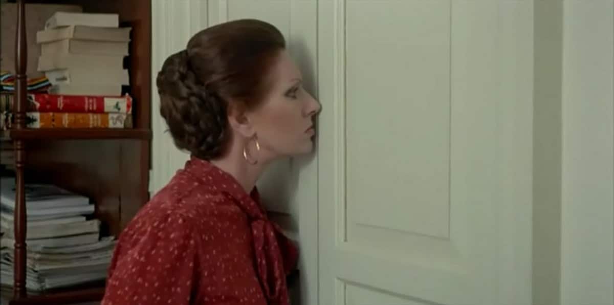 Una mujer con vestido rojo apoya el oído en una puerta cerrada junto a una estantería.