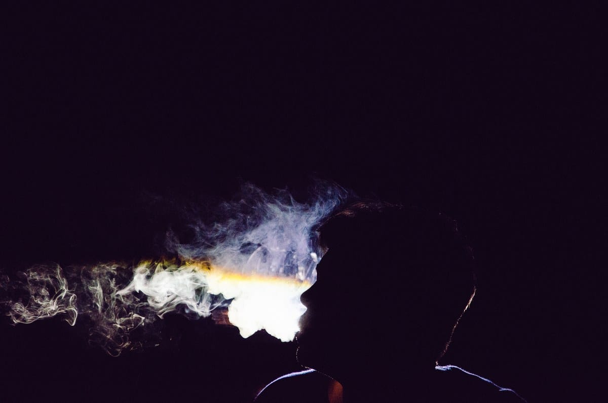 Silueta de una persona de perfil exhalando una densa nube de humo sobre un fondo oscuro