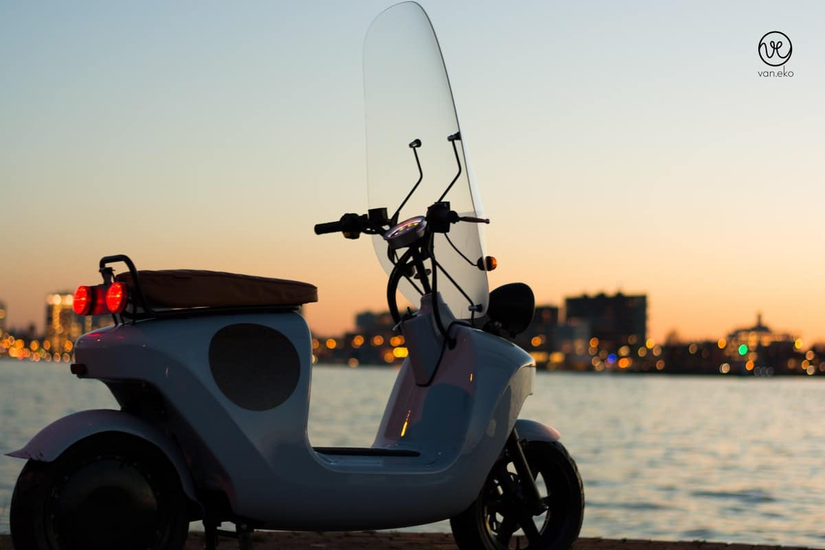 Escúter con parabrisas estacionado junto al agua al atardecer, con luces urbanas al fondo
