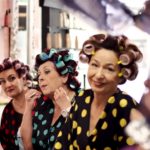 Tres mujeres con batas de lunares y rulos se miran al espejo mientras una estilista trabaja en primer plano.