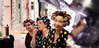Tres mujeres con batas de lunares y rulos se miran al espejo mientras una estilista trabaja en primer plano.