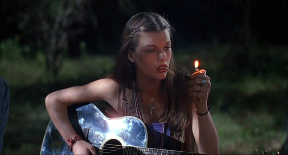 Mujer con una guitarra enciende un cigarrillo al aire libre durante la noche.