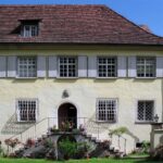 Fachada de una casa histórica con tejado de tejas, ventanas enrejadas y escalinata adornada con flores.