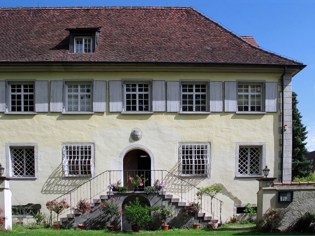 Fachada de una casa histórica con tejado de tejas, ventanas enrejadas y escalinata adornada con flores.