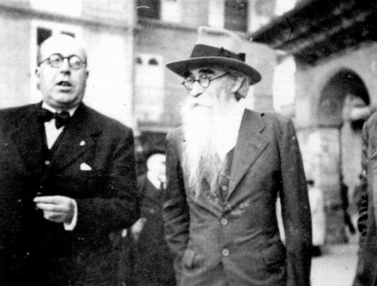 Dos hombres con gafas y traje caminan juntos; uno lleva sombrero y luce una barba larga y blanca