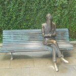 Escultura de bronce de un hombre barbado con gafas, sentado con las piernas cruzadas en un banco