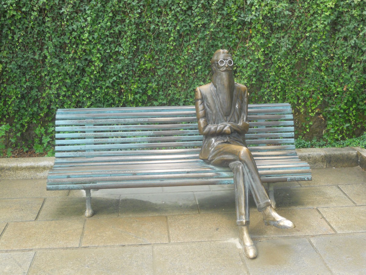 Escultura de bronce de un hombre barbado con gafas, sentado con las piernas cruzadas en un banco