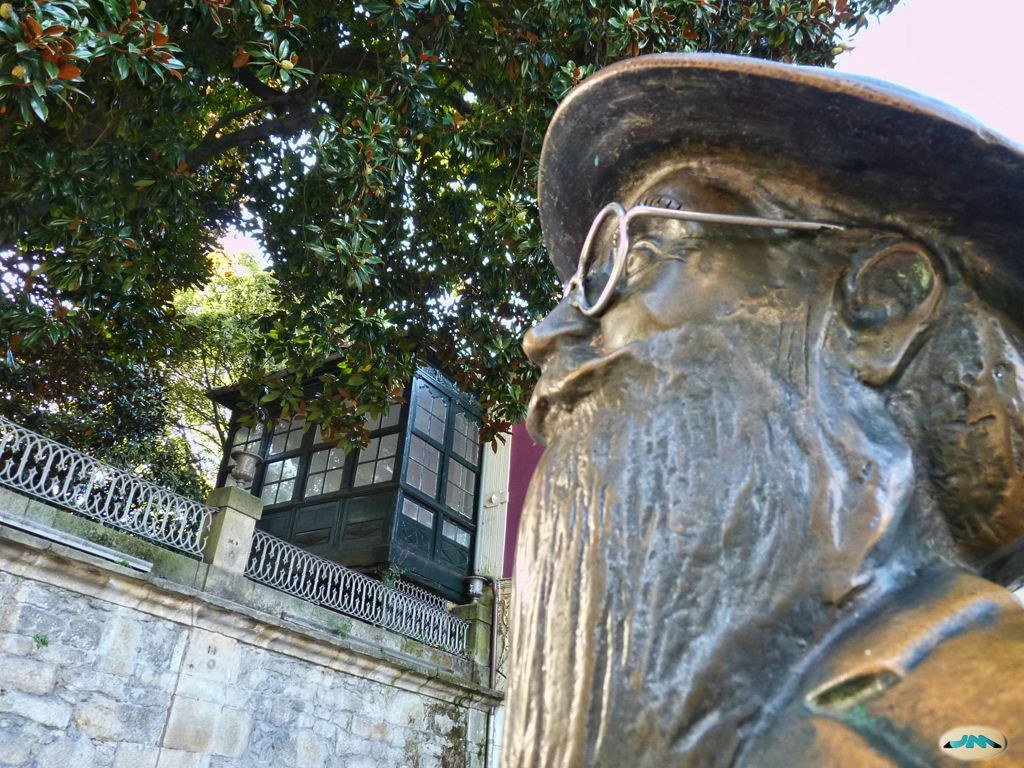 Primer plano de una escultura de bronce con sombrero, gafas redondas y barba larga, ante árboles y un edificio