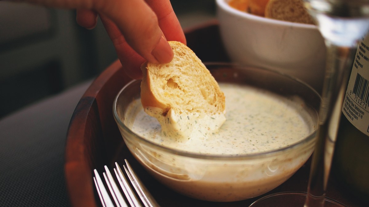 Mano mojando una rebanada de pan en un cuenco de salsa cremosa con hierbas