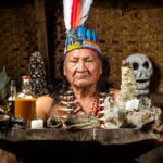 Hombre con tocado ceremonial sostiene una bandeja con velas, frascos, plantas, huesos y un cráneo.