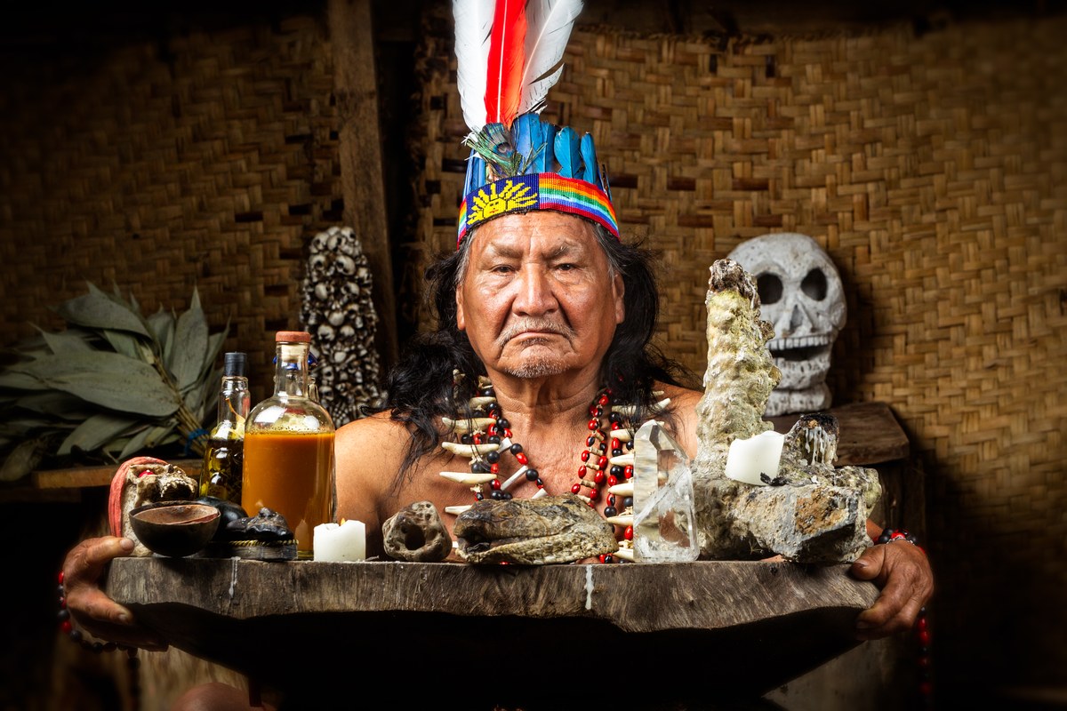 Hombre con tocado ceremonial sostiene una bandeja con velas, frascos, plantas, huesos y un cráneo.