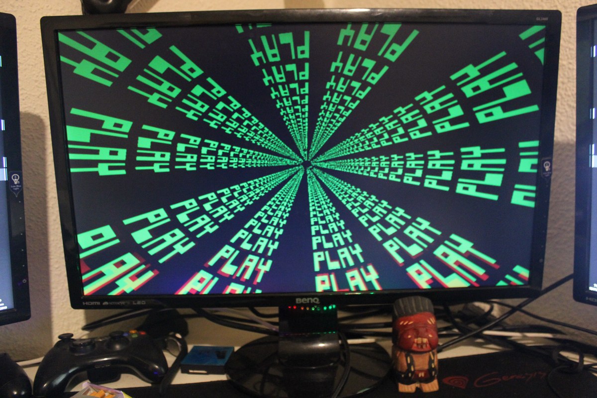 Monitor con la palabra «PLAY» repetida en verde y dispuesta en perspectiva radial; varios mandos reposan delante.