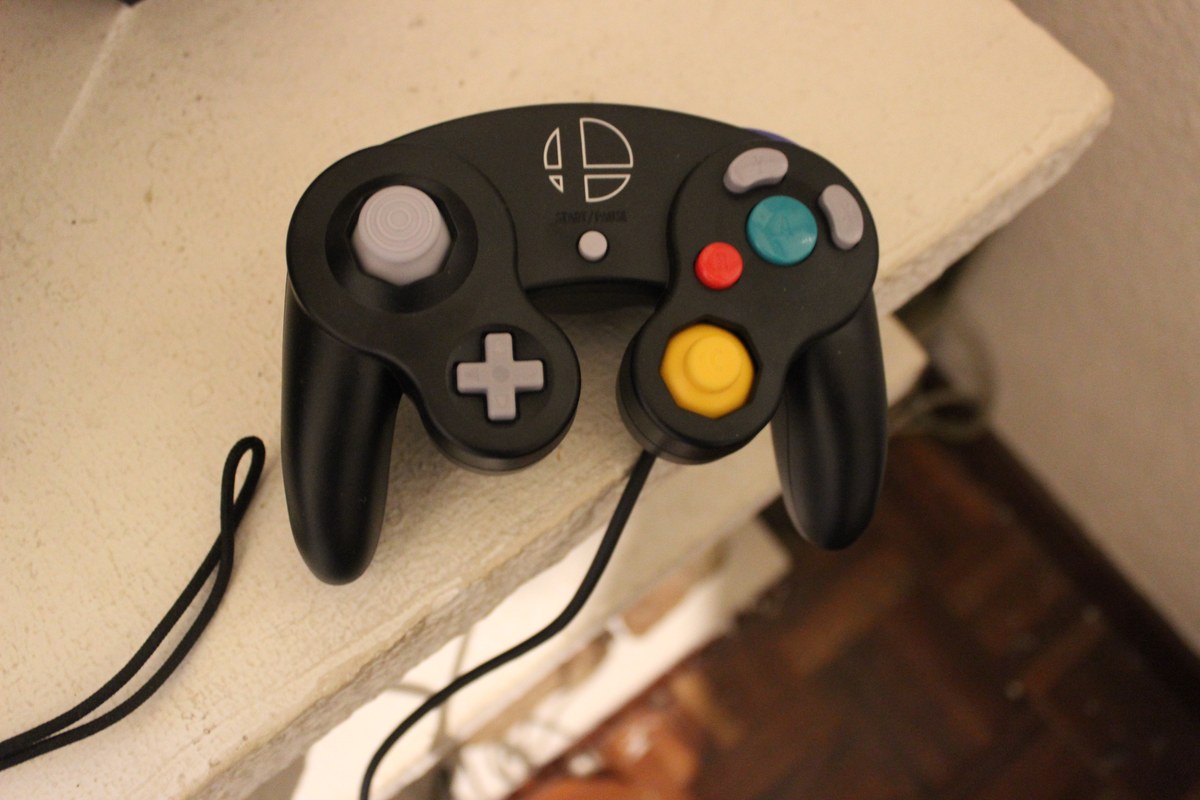 Mando negro con cable, dos palancas, cruceta y botones de varios colores sobre una superficie clara.
