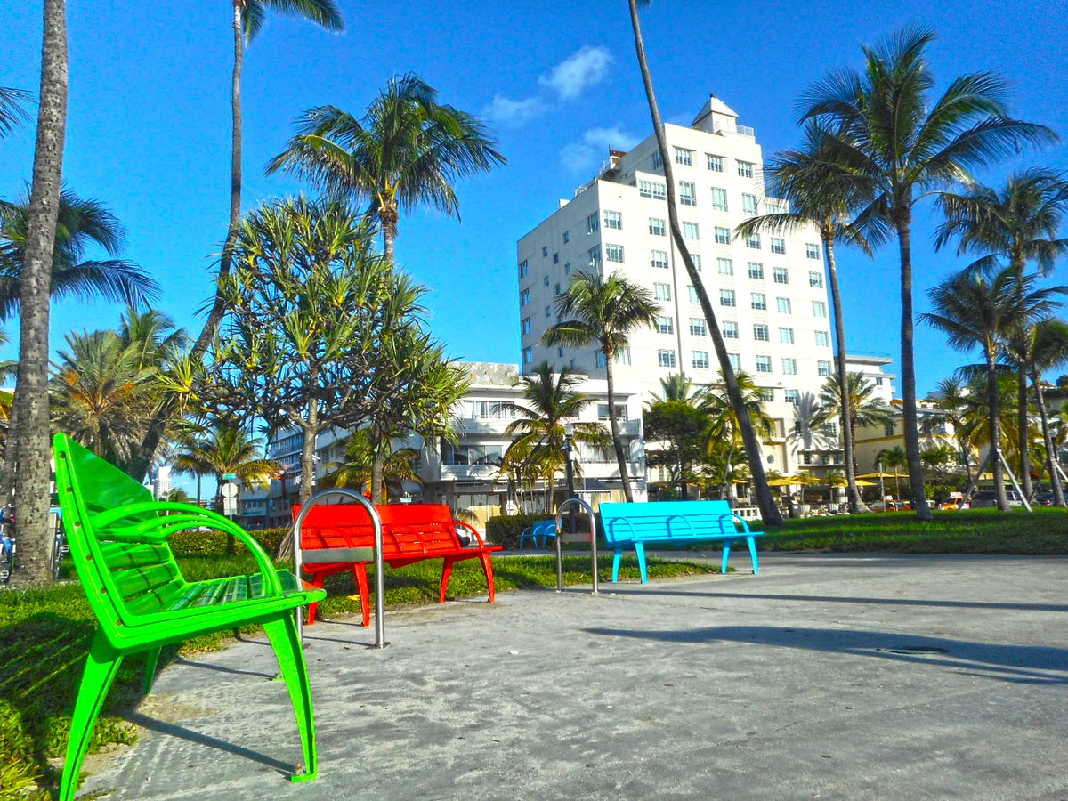 Bancos de colores entre palmeras frente a un edificio blanco de estilo art d&eacute;co