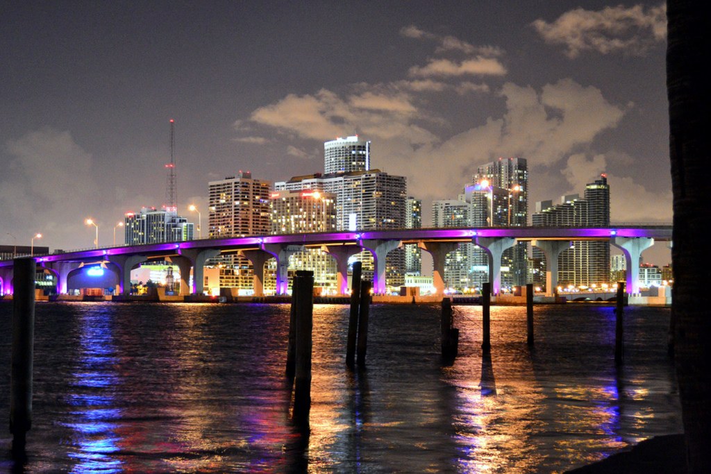 Puente iluminado en violeta ante una ciudad de rascacielos, con reflejos nocturnos sobre el agua