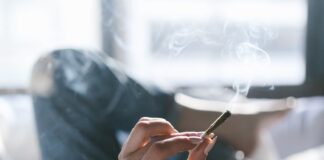 Una persona sostiene un porro encendido mientras permanece tumbada en una cama